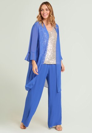 Frau trägt eine blaue transparente lange Jacke und weite Hosen mit einem silbernen Paillettenoberteil und offenen Schuhen, steht und lächelt, heller Hintergrund.