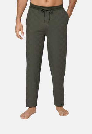 Man draagt donker groene geruite loungebroek met trekkoord, staat blootsvoets met één hand in de zak en de andere ontspannen langs het lichaam.