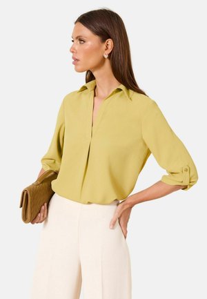 Frau in senfgelber Bluse und cremefarbenen Hosen hält eine braune, geflochtene Clutch, blickt nach links und hat die Hand an der Hüfte vor einfachem Hintergrund.