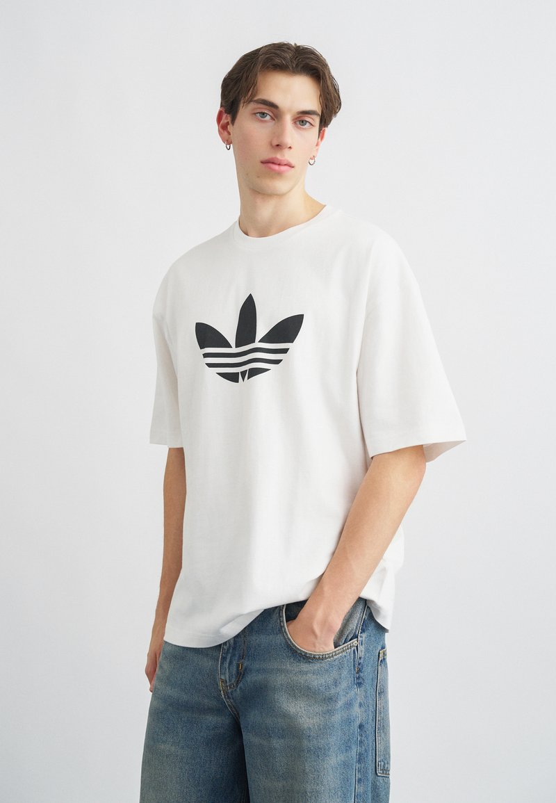 Biele bavlnené tričko s veľkým čiernym logom Adidas trefoil na prednej strane, krátke rukávy a uvoľnený strih. Skombinované s modrými džínsami.