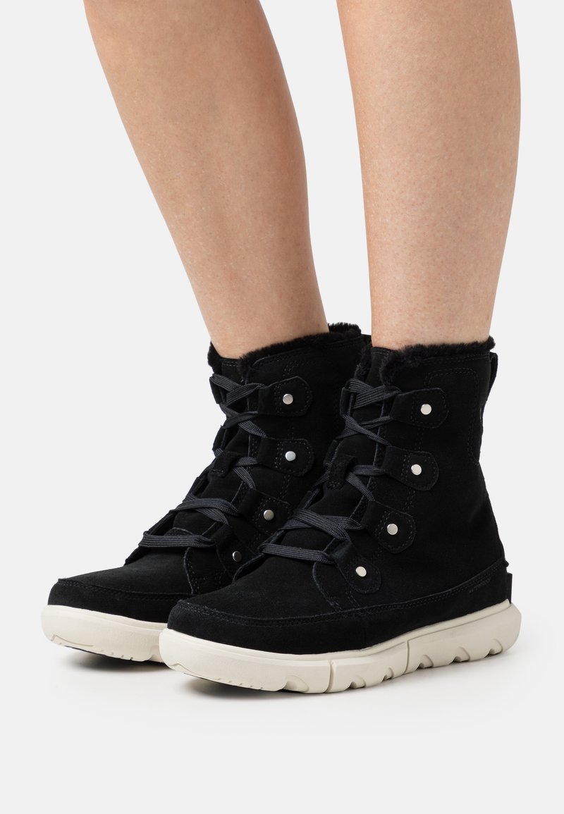 Botas de tobillo de ante negro con forro de piel sintética, presentan acentos plateados, cordones negros y una suela de goma texturizada blanca.