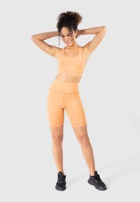 Orange idrotts crop top och matchande högmidjade shorts, tillverkade av elastiskt material med en jämn textur och minimala sömmar, tillsammans med svarta sneakers.