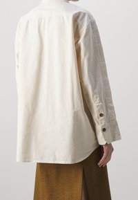 Ljusbeige bomullsskjorta med ryggsyd, långa ärmar, knappade manschetter och rundad nederkant. Bärs över en texturerad brun kjol.