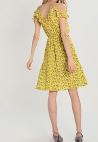 Robe florale jaune avec bretelles à volants, taille élastique et jupe de longueur genoux. Le motif présente de petites fleurs bleues sur un tissu texturé.