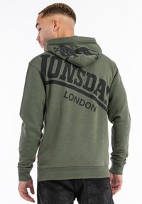 Sudadera de color verde oscuro con un gran logo negro de "LONSDALE" y un gráfico de león en la parte posterior. Tejido suave, puños acanalados y detalle de dobladillo.