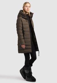 Brun pufferjakke med højt krave, sidelukkere og en skinnende inderside; parret med sorte sneboots med snøring og detaljer.