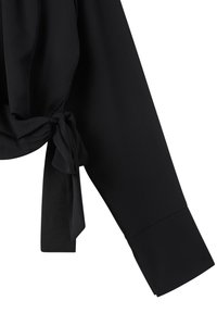Blusa nera con dettaglio nodo legato sul lato sinistro, maniche lunghe e una texture di tessuto liscia e fluida.