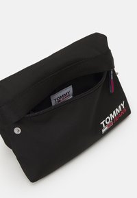 Sacchetto di tessuto nero con design a risvolto, dotato di una tasca con zip e un logo stampato in bianco e rosso. Superficie testurizzata e dettaglio di chiusura in metallo.