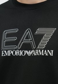 Fekete pulóver fehér "EA7 EMPORIO ARMANI" felirattal, modern betűtípussal. Puha tapintású anyag, bordázott nyakrésszel és logó részletekkel.