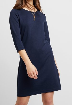 Frau trägt ein marineblaues, knielanges Kleid mit dreiviertellangen Ärmeln und eine goldene Anhängerhalskette, steht vor einem schlichten Hintergrund.