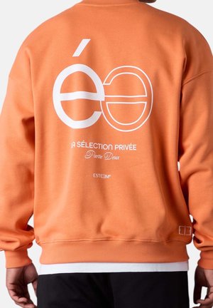 Persona con una sudadera naranja con texto gráfico blanco y símbolos en la espalda, de pie frente a un fondo claro y sencillo.