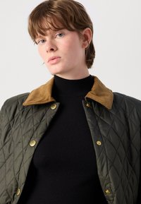 Gev quilted olijfgroene jas met een bruine corduroy kraag, voorzien van gouden drukknopen en een strak zwart trui eronder.