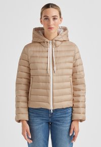 Beige Steppjacke mit Kapuze, mit horizontalem Steppmuster, weißem Reißverschluss und Zugbändern. Über Jeans getragen.