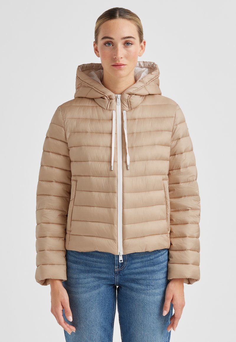 Beige Steppjacke mit Kapuze, mit horizontalem Steppmuster, weißem Reißverschluss und Zugbändern. Über Jeans getragen.