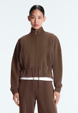 Vrouw draagt een bruine jas met rits en koord in de taille en bijpassende broek met verticale naaddetails, staand tegen een effen achtergrond.