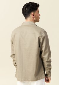 Ljusbeige knapp-up jacka med krage, långa ärmar och manschetter med knappar. Slät textur och enkel design med minimala detaljer.