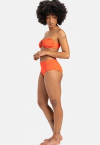 Oranje bandeau bikini met geribbelde textuur en zwarte hardwaredetails. Hooggeplaatste onderkant. Model poseert zijwaarts op een neutrale achtergrond.