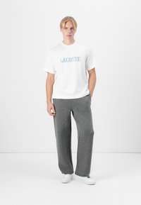 Biała bawełniana koszulka z nadrukiem "LACOSTE" w niebieskim i zielonym kolorze, w połączeniu z szarymi, luźnymi, teksturowanymi spodniami oraz białymi sneakersami.