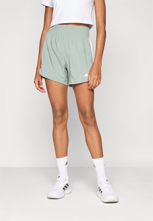 Femme portant un short de sport Adidas vert clair, un haut court blanc, des chaussettes Adidas blanches et des baskets Adidas blanches, debout devant un fond gris.