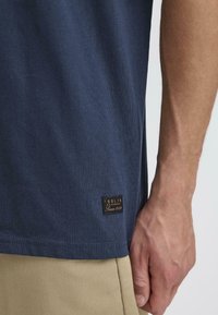 Tricou din bumbac bleumarin cu mânecă scurtă, având un eticheta cusută de culoare maro la margine. Materialul are un finisaj neted, ușor texturat.