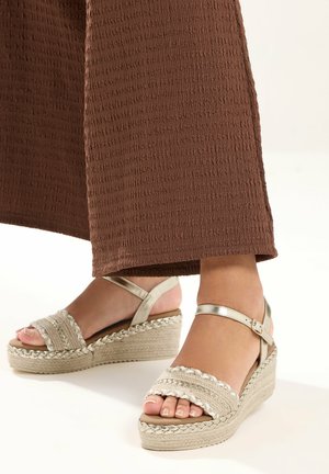 Voeten met beige gevlochten sleehakken met enkelbandjes, gecombineerd met bruine, getextureerde broek met wijde pijpen op een witte achtergrond.