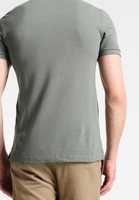 Chemise verte à manches courtes avec un tissu texturé, coupe ajustée et ourlet droit, portée avec un pantalon beige, vue de dos.