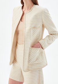 Kremowo-beżowa marynarka w prążki z okrągłym dekoltem, przednią kieszenią i plecionymi wykończeniami, w zestawie z dopasowanymi szortami.