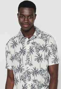 Camicia a maniche corte con bottoni in tessuto leggero, caratterizzata da uno sfondo bianco con motivi di palme scure e bottoni di legno.