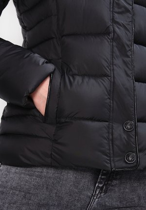 Ręka w kieszeni czarnej pikowanej kurtki z zatrzaskami, zestawiona z ciemnoszarymi dżinsami, przedstawiona od talii do połowy uda.