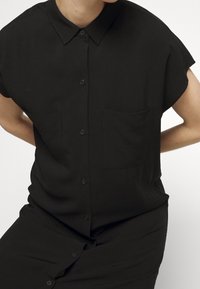 Black short-sleeve shirt with a button-up front, two chest pockets, and a classic collar; smooth fabric with no visible patterns or designs.
