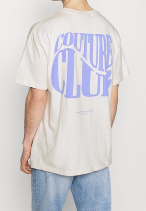 T-shirt oversize beige avec grand texte violet "COUTURE CLUB" au dos, en coton, manches courtes, coupe décontractée.