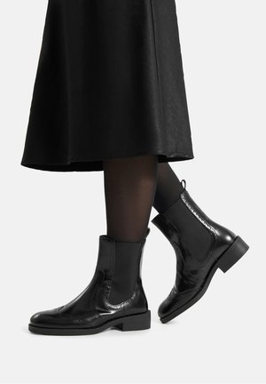 Czarne błyszczące botki za kostkę z elastycznymi panelami po bokach, zdobione ażurowym wzorem typu brogue, na niskim, szerokim obcasie. Noszone z czarnymi rajstopami i czarną spódnicą.