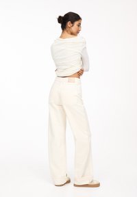 Jean à jambes larges de couleur claire avec une texture lisse, comportant une poche arrière et une petite étiquette de marque sur la taille, associé à un haut fluide.