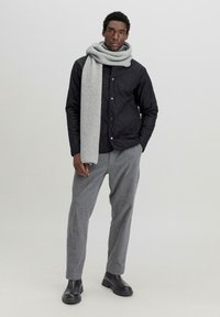 Schwarze gesteppte Jacke mit grauem geripptem Schal, grauen maßgeschneiderten Hosen und schwarzen Stiefeletten, vor einem schlichten hellen Hintergrund präsentiert.