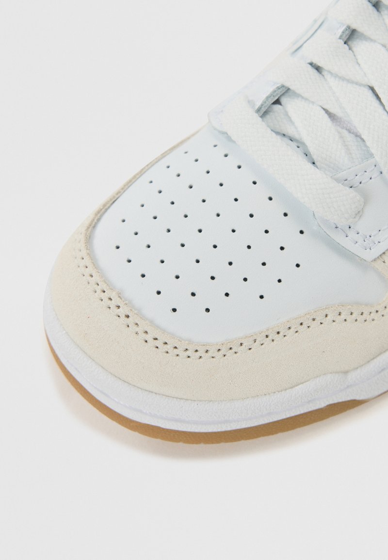 Zapatilla blanca con parte superior de cuero suave y acentos de ante. Caja de puntera perforada, cordones blancos planos y suela de goma natural.