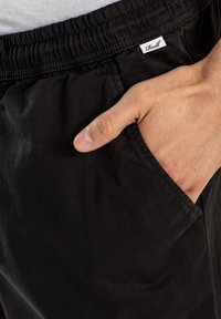 Pantalones de algodón negros con una cinturilla elástica. Una mano descansa en el bolsillo lateral, mostrando la textura suave de la tela y un ligero brillo.