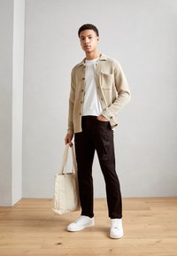 Cardigan beige texturé avec des boutons, T-shirt blanc, pantalons noirs déchirés et baskets blanches. Tient un sac fourre-tout beige. Fond de sol en bois.