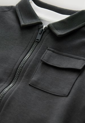 Sort zip-up jakke med krave, featuring en frontlomme og tekstureret stof. Lynlåsen er sort, og der er en hvid foring indeni.