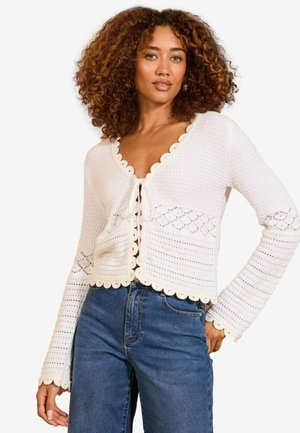 Weißer Häkel-Cardigan mit gewellten Kanten, V-Ausschnitt, Bindebänder und langen Ärmeln. Über blauen Jeans getragen.