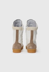 Un paio di sneakers alte bianche e beige in suede con suole in gomma, viste da dietro su uno sfondo bianco semplice.