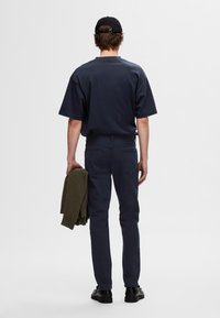 Laivastonsininen ylipitkä T-paita, tummansiniset slim-fit -housut, mustat kengät, käsissään teksturoitu oliivinvihreä neule. Minimalistinen muotoilu, ei näkyvää brändäystä.