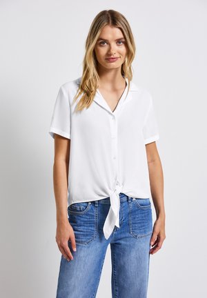 Camisa blanca de manga corta con cuello y detalle de lazo en la parte delantera, hecha de una tela suave, combinada con jeans de mezclilla azul claro.