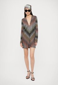 Minišaty s dlhými rukávmi s farebným cikcak vzorom, hlbokým véčkovým výstrihom, priliehavým strihom a trblietavým pleteným materiálom. Nosia sa s čiernymi lodičkami.