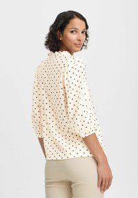 Gräddfärgad blus med bruna prickar, textil med struktur, puffärmar och flätad krage. Bärs tillsammans med beige byxor, visande bakifrån.