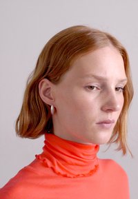 Un col roulé rouge côtelé avec un col volanté ; cheveux châtain ondulés et courts ; portant des boucles d'oreilles en argent. Éclairage doux et fond neutre.