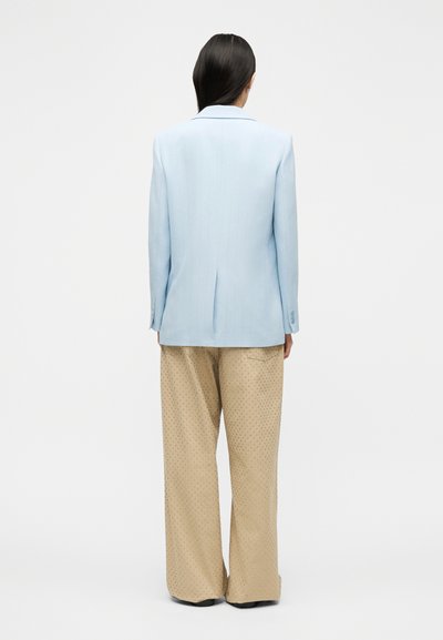 Γυναίκα με μακριά σκούρα μαλλιά, φορώντας γαλάζιο σακάκι και μπεζ παντελόνι φαρδύ στο κάτω μέρος με μικρές τρουκς, στέκεται πλάτη σε απλό φόντο.
