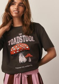 Brun t-shirt med et grafisk design, der viser røde svampe med teksten "THE TOADSTOOL" ovenfor og "Natures tiny umberella" nedenfor.