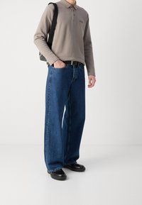 Man wearing taupe long-sleeve polo, loose blue jeans, black belt, black shoes, and carrying black shoulder bag on plain white background.