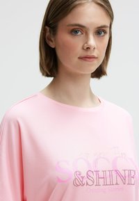 Young woman with short brown hair wears a light pink shirt with pink and rhinestone text reading "SHINE" on a plain background.
