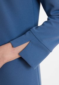 Manche en tissu bleu avec une finition texturée, présentant un détail découpé au niveau du poignet. Gros plan montrant l'emplacement de la main contre le tissu.